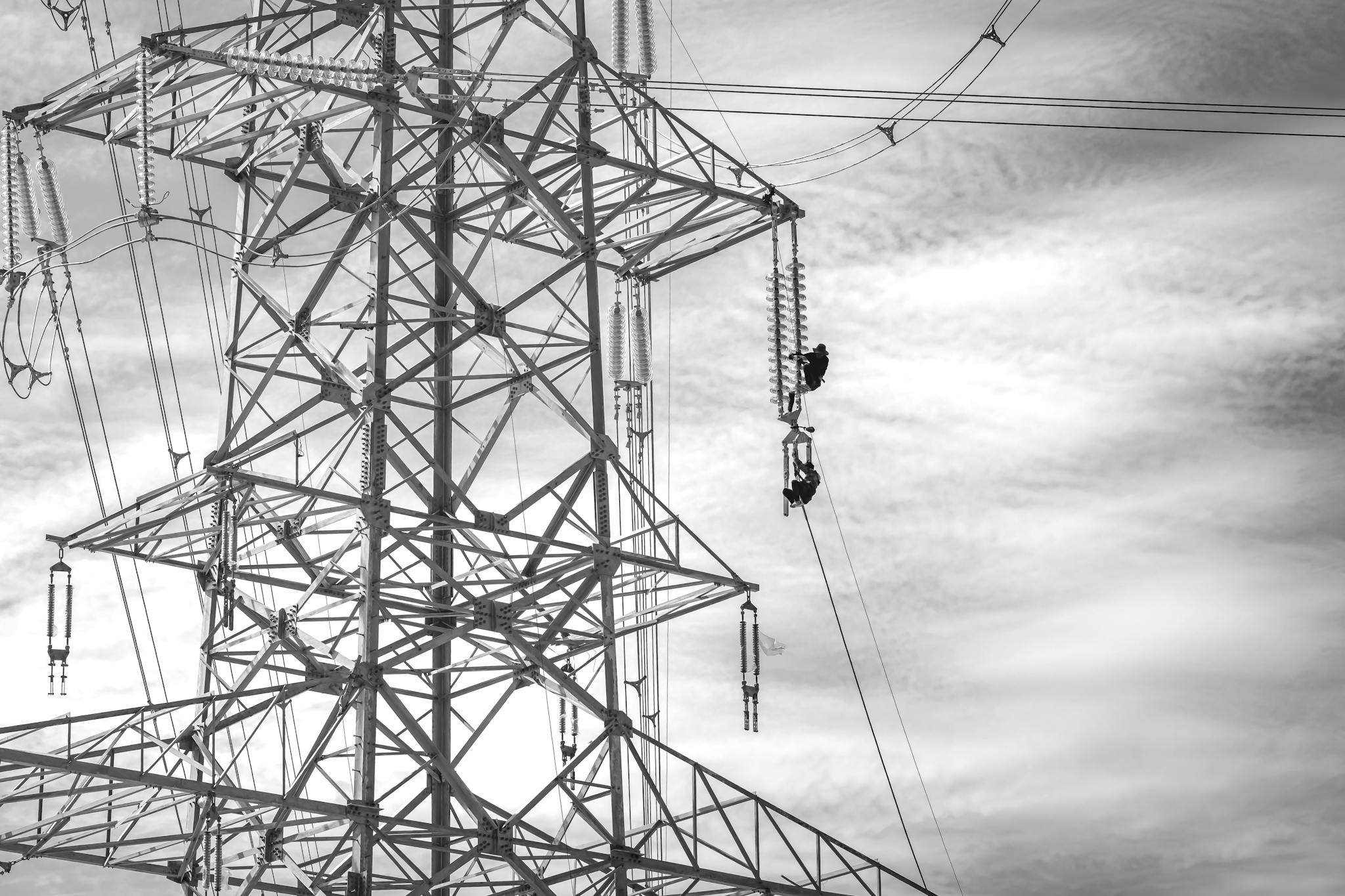 Black and white image of workers performing maintenance on a transmission tower against a cloudy sky.